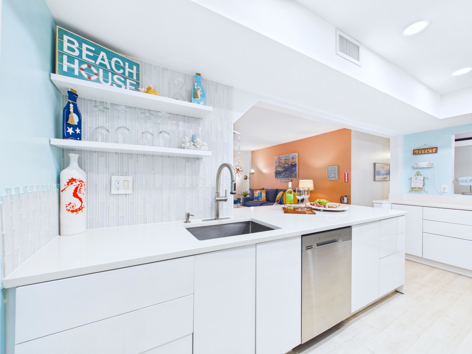 A modern kitchen with white cabinetry, a stainless steel sink, and a built-in dishwasher. There are decorative items on the shelves, including a blue bottle and a glass vase.