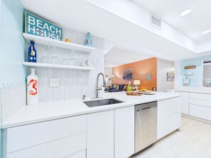 Une cuisine moderne avec des meubles blancs, un évier en acier inoxydable et un lave-vaisselle encastré. On peut y voir des objets décoratifs sur les étagères, notamment une bouteille bleue et un vase en verre.