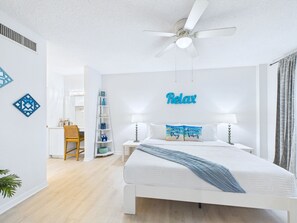 Une chambre à coucher comprenant un lit, un bureau, une chaise, un ventilateur de plafond et un panneau mural sur lequel est écrit « Détendez-vous ».