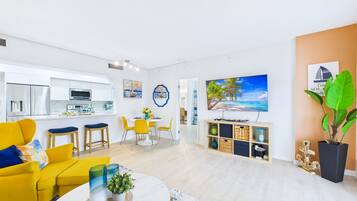 Een moderne woonkamer met een gele bank, een eethoek met gele stoelen, een keuken met witte kasten, en een groot schilderij van een strandlandschap.