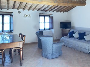 A living room with a wooden dining table, chairs, a patterned sofa, a television, and a window with curtains. - Camigliano Wine State - Ghiandaie (Camigliano)