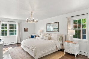 A bedroom with a large bed, two bedside tables, a chandelier, and a window with curtains. - Pondside Home | Hot Tub • Fire Pit • Beach Access (Greenwood Lake)