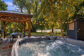 A hot tub with a pergola and string lights, surrounded by a patio area with chairs and a table. - Pondside Home | Hot Tub • Fire Pit • Beach Access (Greenwood Lake)