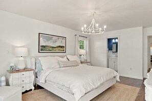 A bedroom with a large bed, bedside tables, a chandelier, a window with curtains, and a painting on the wall. - Pondside Home | Hot Tub • Fire Pit • Beach Access (Greenwood Lake)
