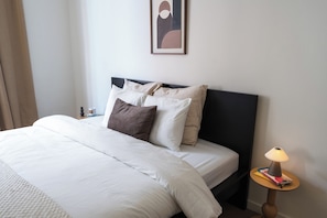 En snyggt bäddad säng med vita sängkläder och flera kuddar, ett sängbord med en lampa och ett inramat, abstrakt konstverk på väggen.