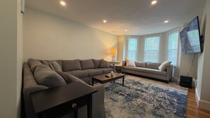 Uma sala de estar com um sofá seccional, um sofá menor, uma mesa de centro e uma TV fixada na parede.