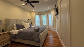 Quarto com cama, mesa de cabeceira, luminária, ventilador de teto e janelas com persianas.