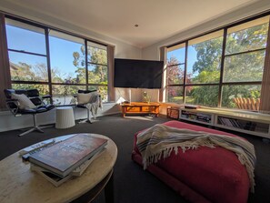 A modern living room with a large window, a flat-screen TV, a red sofa, a coffee table with books, and a chair with a cushion. - Makers Rest a relaxing 4BR 3Bath for 6 with its own Poolfresh Wellness Retreat (Wynyard)