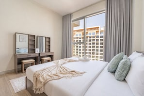 A hotel room with a bed, a desk with a chair, a small stool, a window with curtains, and a view of buildings outside. - Stunning Gulf of Oman Views, 2BR with Assistant Room, Private Beach Access (Khor Fakkan)