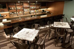 Zona de bar con una barra larga, varios estantes llenos de botellas y mesas con sillas.