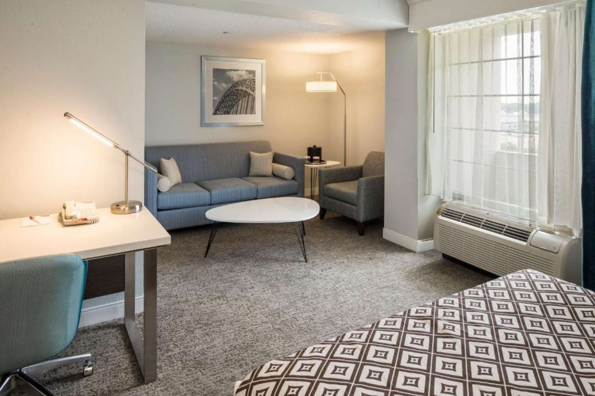 Habitación de hotel con cama, escritorio, sofá, sillón, mesita, lámpara, radiador y ventana con cortinas.