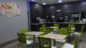 A modern dining area with green chairs and white tables, a kitchen area with appliances, and a TV screen displaying a news program. - Downtown Houston + Free Breakfast & Parking (Houston)