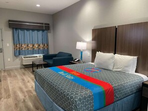 Una habitación de hotel con cama, escritorio, silla, lámpara y ventana con cortinas.