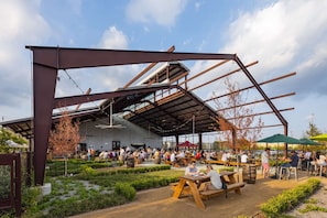 Un área de comedor al aire libre con mesas y bancos, rodeada de follaje y bajo una estructura metálica grande.