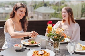 Deux personnes sont assises à une table à l’extérieur, devant une variété d’aliments pour le petit-déjeuner, notamment des gaufres, une tasse de café et un verre de jus d’orange.