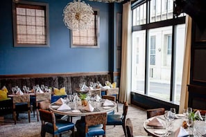 Un restaurant avec des tables rondes dressées pour le dîner, un grand lustre et une vue sur la rue grâce à de grandes fenêtres.
