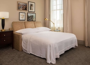 A neatly made bed with a white comforter and pillows, a bedside table with a lamp, and a small round table. - 1BR King Steps to James River + Dining (Lynchburg)