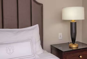 A neatly made bed with a pillow featuring a crest emblem, a bedside table with a lamp, and a wall socket. - 1BR King Steps to James River + Dining (Lynchburg)