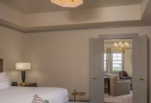 Habitación de hotel con cama, mesita de noche, lámpara y vista a un comedor por una puerta abierta.