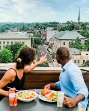 Two people dining at a table with a view of a cityscape, including buildings and a church spire. - 1BR King Steps to James River + Dining (Lynchburg)
