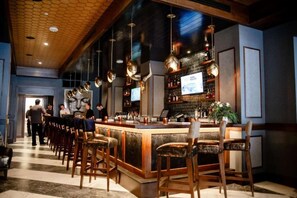 A bar area with wooden bar stools, a counter with a television, and shelves with bottles. - Stay Lynchburg Iconic Building + Rooftop Dining (Lynchburg)