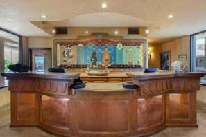 Un mostrador de recepción con un mural decorativo, un letrero que indica un restaurante y una zona de recepción con piso de baldosas.