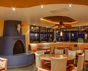 Ein Restaurant mit runden Tischen, Stühlen, einem Kamin und Blick auf die beleuchtete Stadt.