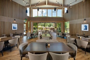 Un'ampia sala da pranzo interna con diversi tavoli e sedie, ampie finestre che offrono vista sull'esterno e un moderno design del soffitto con lampade a sospensione.