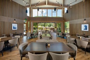 Un'ampia sala da pranzo interna con diversi tavoli e sedie, ampie finestre che offrono vista sull'esterno e un moderno design del soffitto con lampade a sospensione.