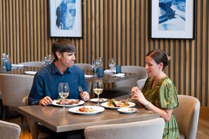 A couple dining at a restaurant with a modern interior, featuring wooden paneling and framed artwork on the walls. - 3BR w/Kitchen & Balcony + 2 Heated Pools & Town Shuttle (Vail)