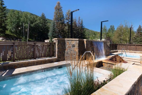 A hot tub area with a waterfall feature, surrounded by stone walls and a wooden fence. - 3BR w/Kitchen & Balcony + 2 Heated Pools & Town Shuttle (Vail)