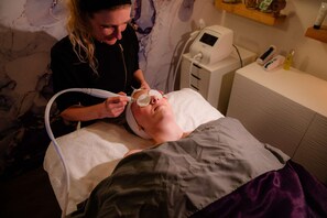 Una persona sdraiata su un letto riceve un trattamento viso, con un apparecchio medico e una bottiglia su un ripiano nelle vicinanze.