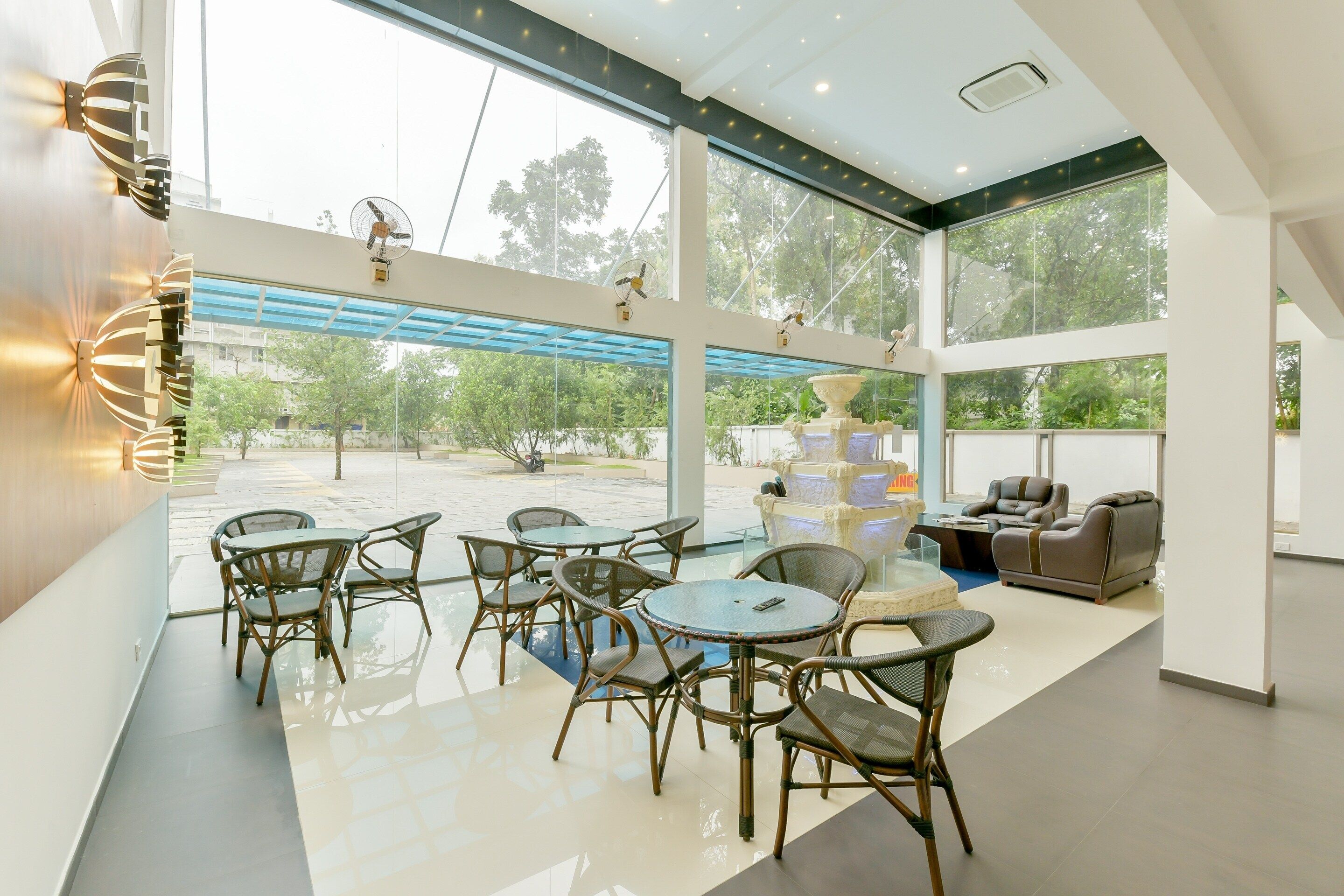 A modern indoor space with a dining area featuring round tables and chairs, a seating area with sofas and armchairs, and large windows offering views of the outdoors.