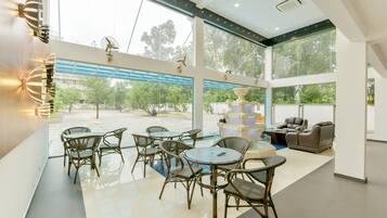 A modern indoor space with a dining area featuring round tables and chairs, a seating area with sofas and armchairs, and large windows offering views of the outdoors.