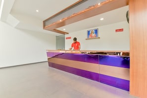 A reception desk with a person standing behind it, a framed picture on the wall, and a clock indicating 12 PM. - Super Townhouse Edassery Blue Bells (Kochi)