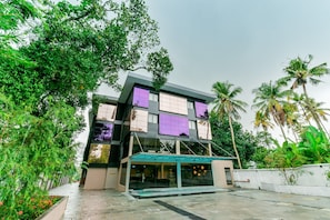 A modern multi-story building with a flat roof, surrounded by lush greenery and palm trees. - Super Townhouse Edassery Blue Bells (Kochi)