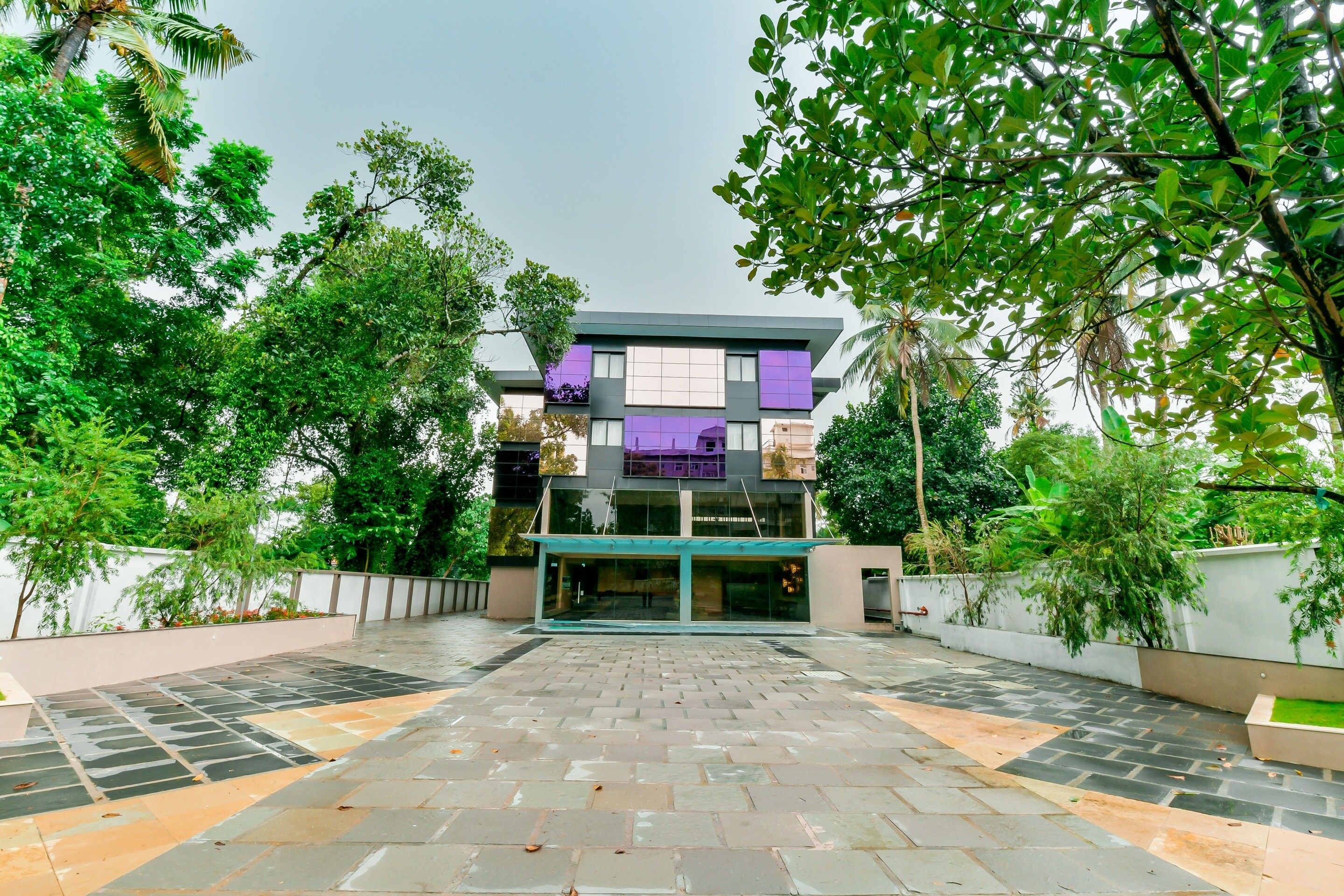 A modern building with a glass entrance, surrounded by a paved courtyard and lush greenery.