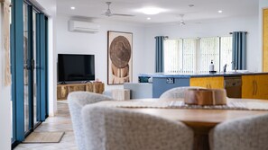 A modern living room with a flat-screen TV, a dining area with a wooden table, and a kitchen with a blue countertop. - Wongaling Whispers~ Escape to the gentle rhythm of Wongaling Beach (Wongaling Beach)