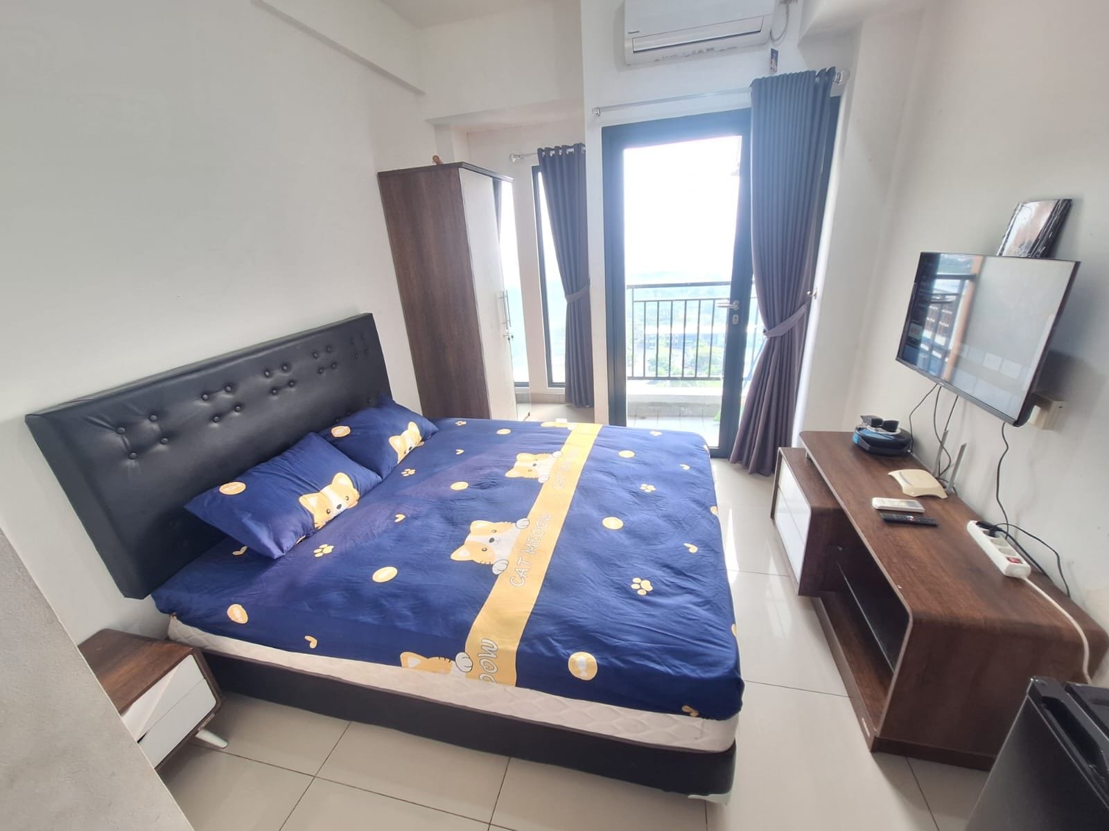 A bedroom with a bed, a bedside table, a TV, and a window with curtains.