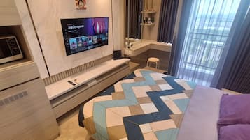 A modern hotel room with a flat-screen TV, a microwave, a vanity with a mirror, a bed with a patterned bedspread, and a window with curtains.