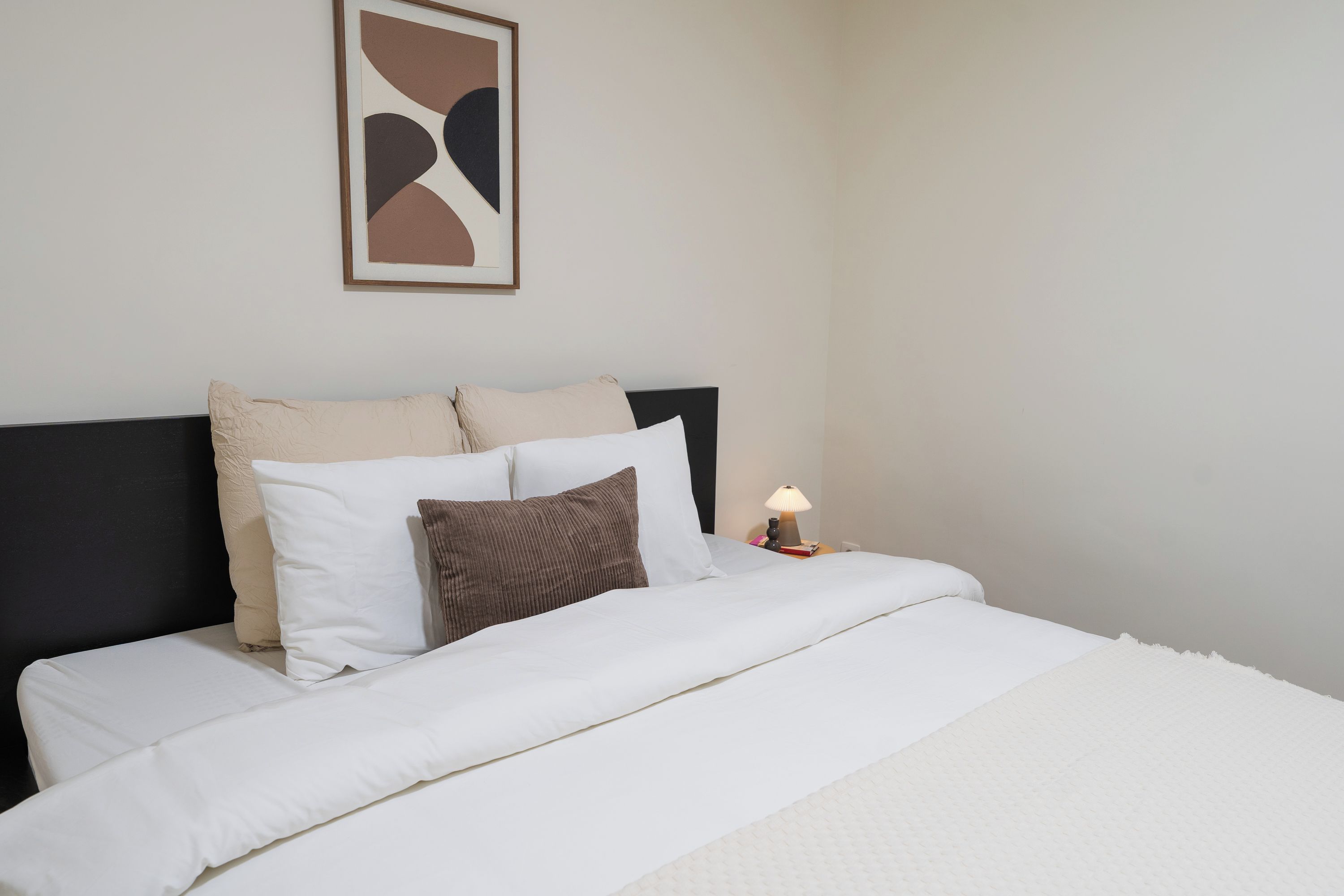A neatly made bed with white and brown bedding, a framed abstract artwork on the wall, and a bedside table with a lamp.