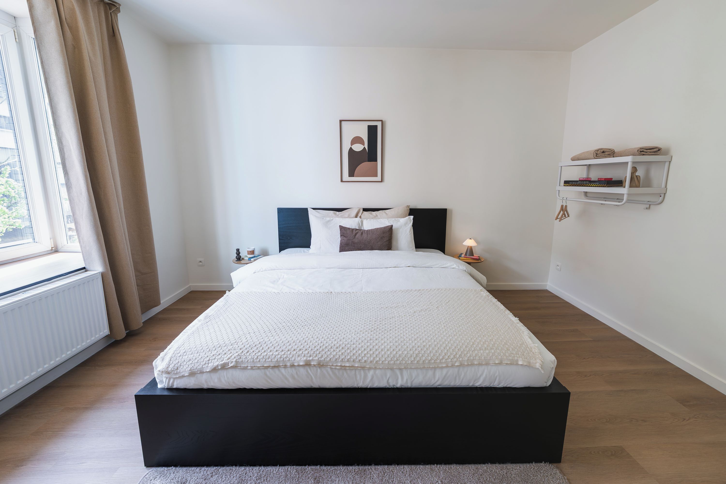A bedroom with a bed, a window with curtains, a bedside table, a wall-mounted shelf, and a framed picture.