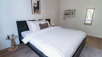 Una cama bien hecha con cabecera, una mesita de noche con lámpara, un cuadro en la pared y una estantería con objetos.