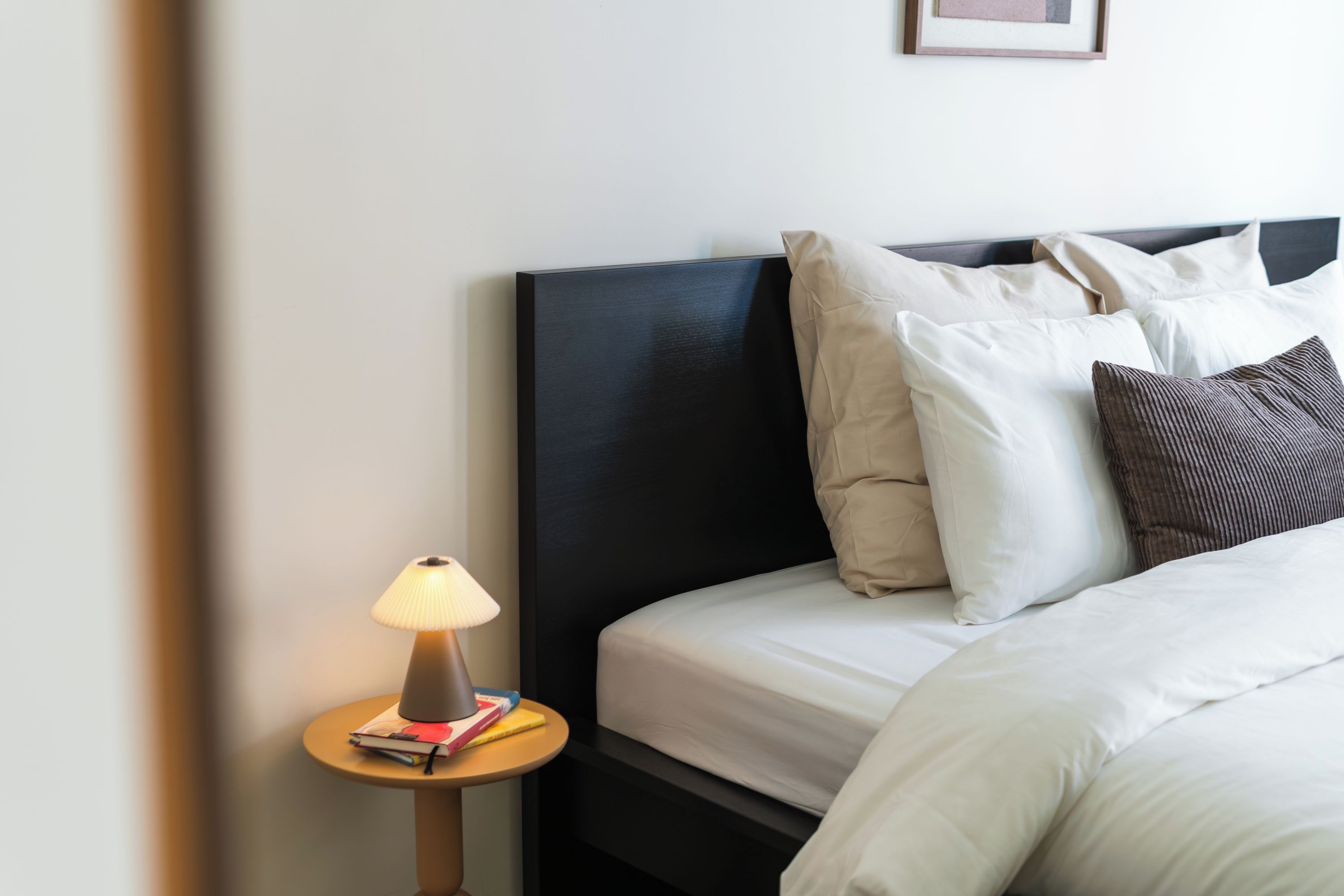 Una cama bien hecha con ropa blanca y varias almohadas, una mesita de noche con una lámpara y un cuadro en la pared.