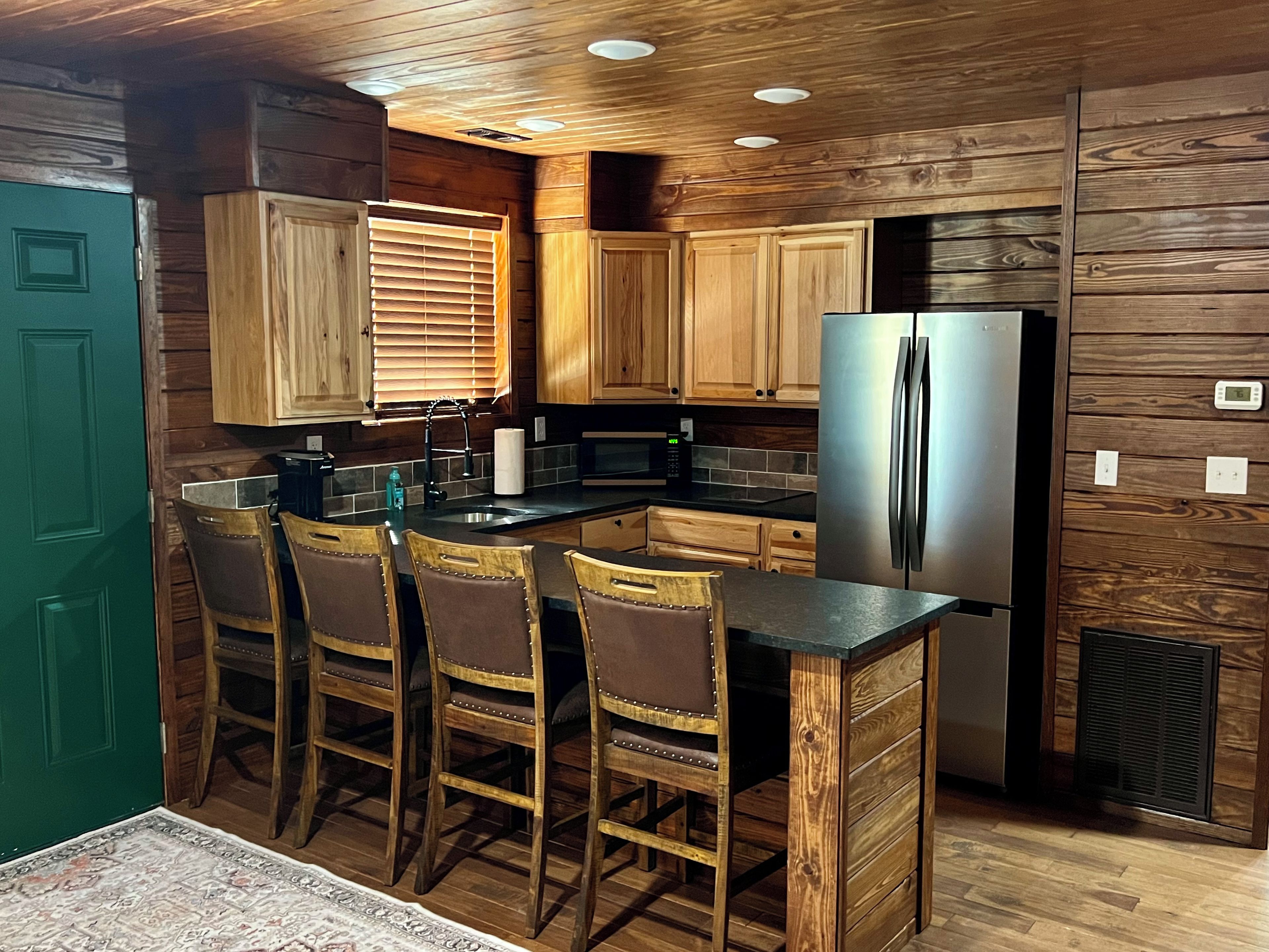A compact kitchen with wooden cabinetry, a stainless steel refrigerator, and a bar area with stools.