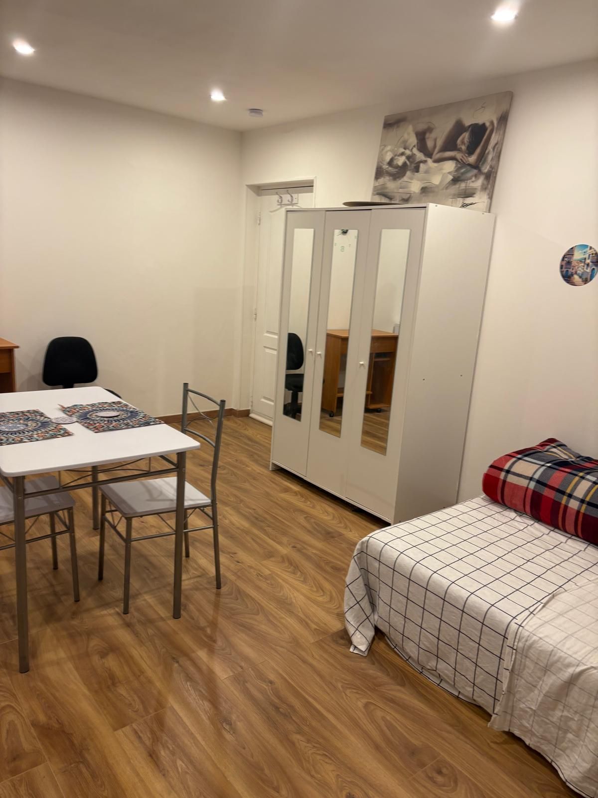 Een kamer met een eettafel, stoelen, een garderobe en een bed met een geruit plaid.