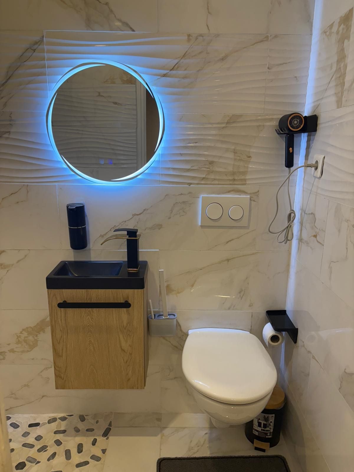 A bathroom with a toilet, a sink with a wooden cabinet, a mirror with a blue light, and a handheld showerhead.