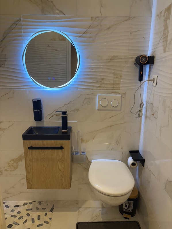A bathroom with a toilet, a sink with a wooden cabinet, a mirror with a blue light, and a handheld showerhead. - Vrbo Property (Lille)