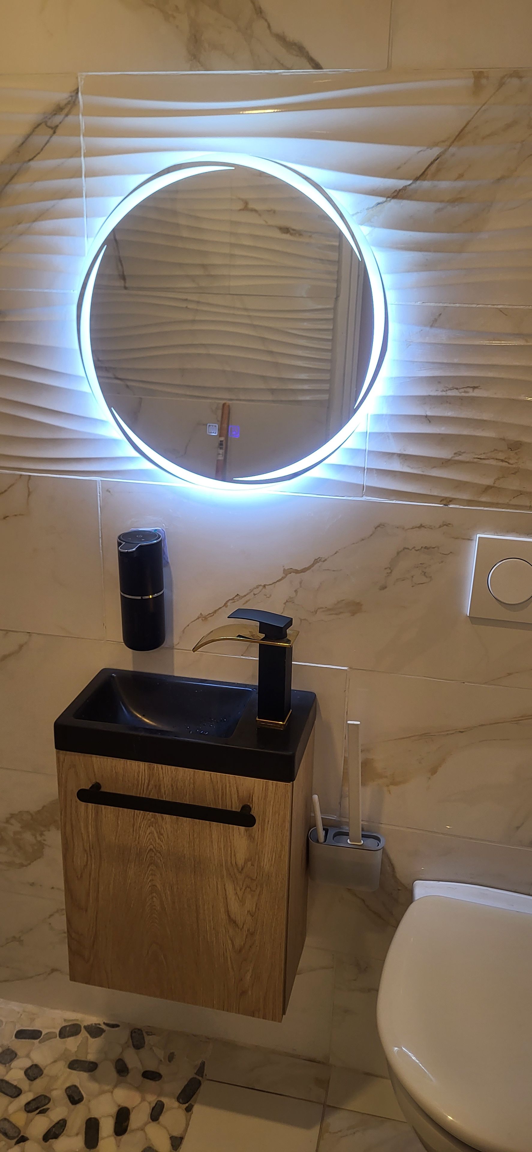 A bathroom with a wooden vanity, a sink, a toilet, and a wall-mounted mirror with a circular light.