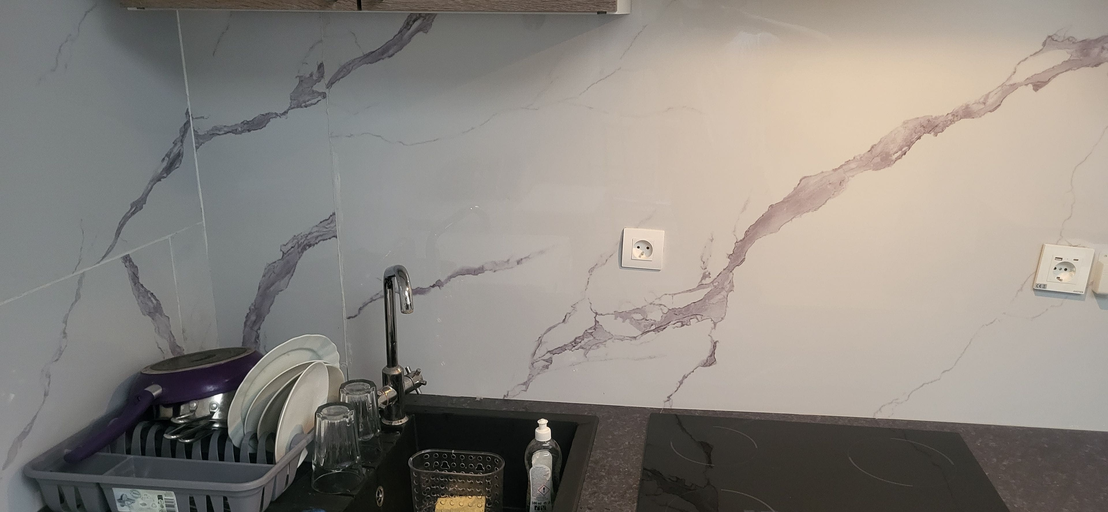 A kitchen with a marble backsplash, a dish rack, a sink, and a bottle of hand soap.